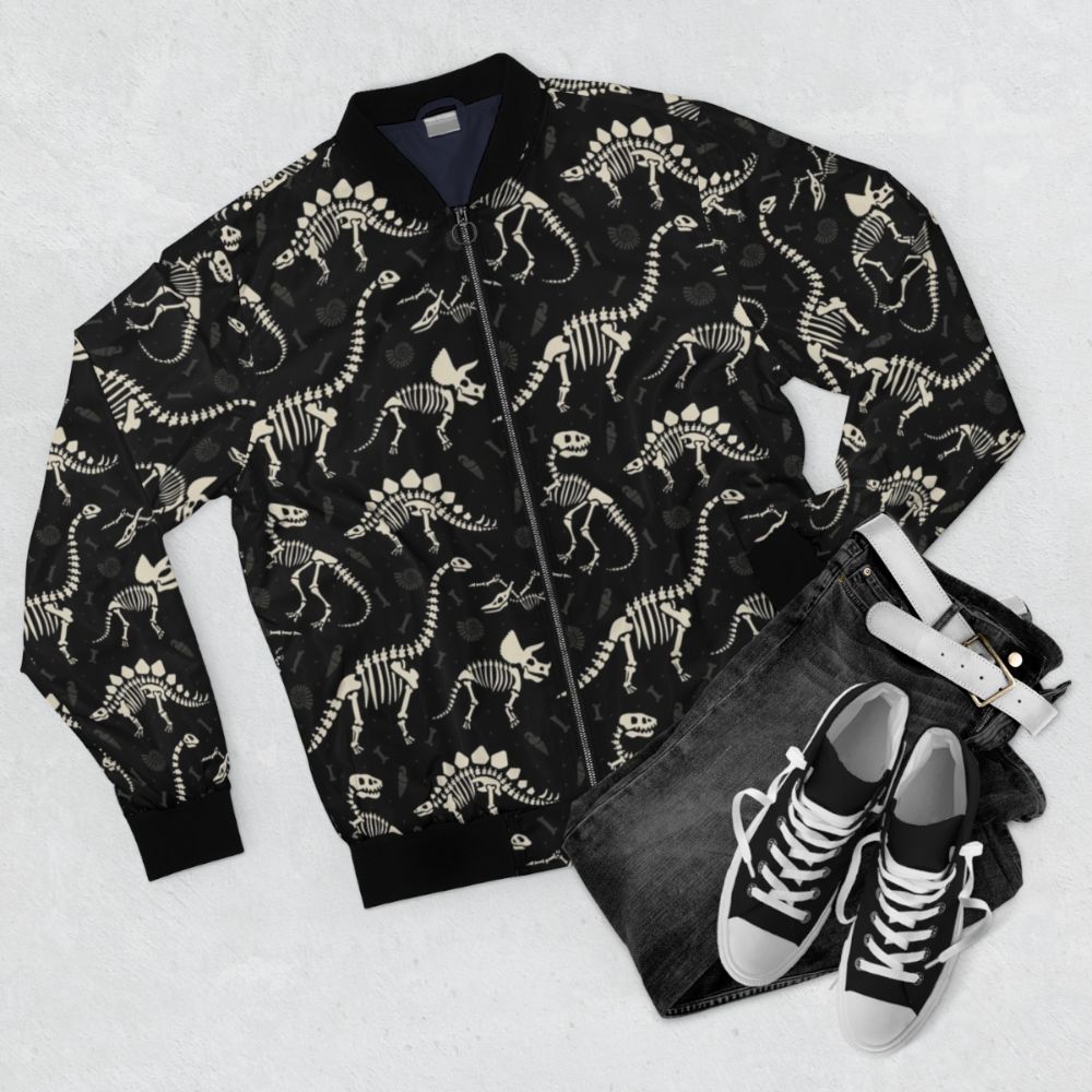 Black bomber jacket with a dinosaur fossil print design, featuring T-Rex, Triceratops, and other prehistoric creatures. - Flat lay