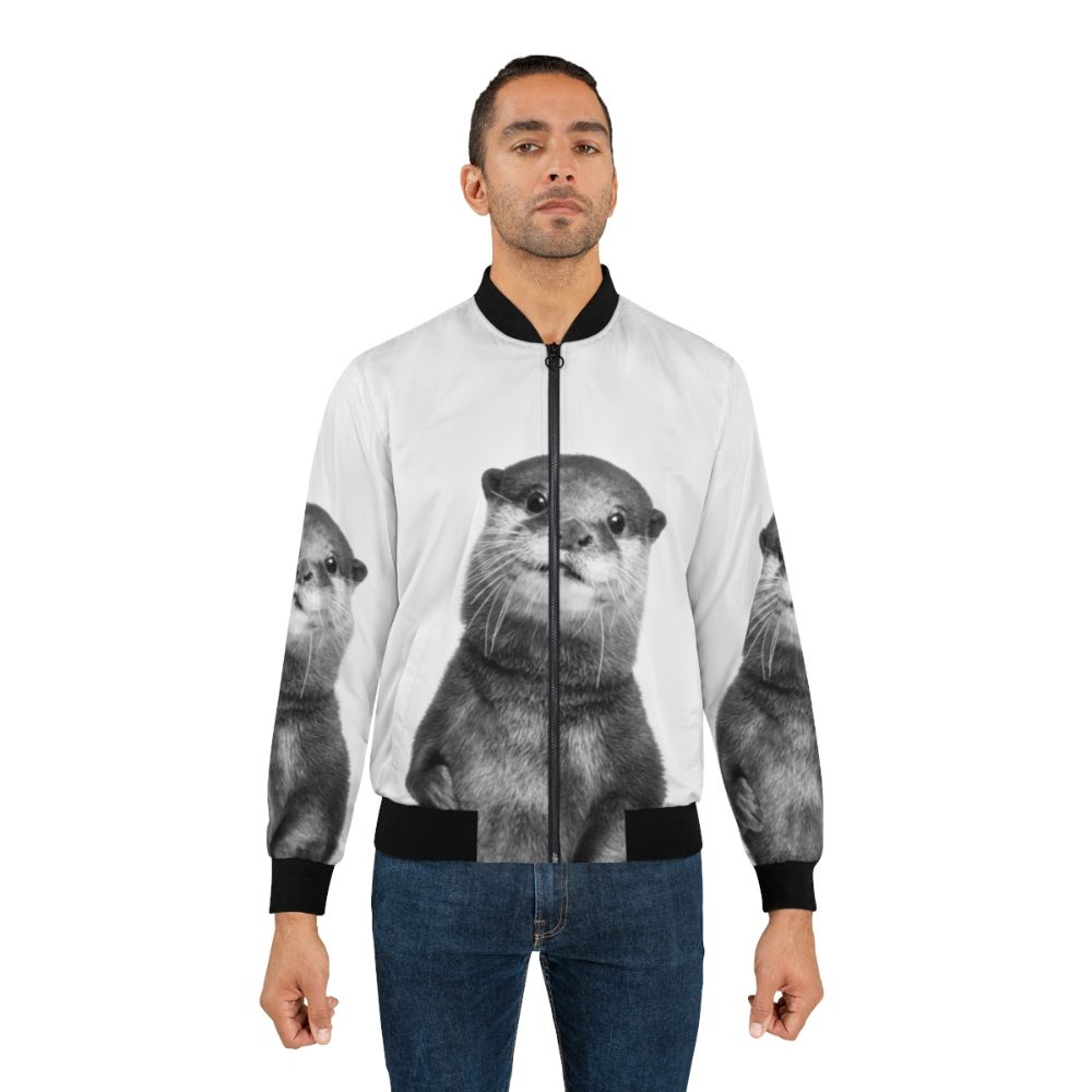 A black and white photo of a smiling otter wearing a bomber jacket, surrounded by a forest and water. - Lifestyle