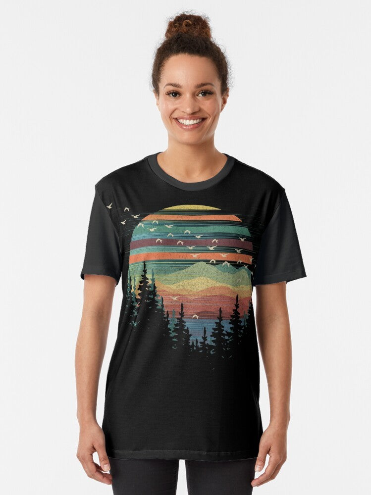 A graphic t-shirt featuring a geometric nature sunset vibes design with elements like forests, mountains, birds, and the sky. - Women