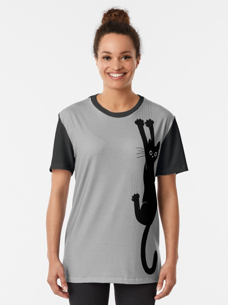 Graphic t-shirt featuring a black cat holding on with its claws and an expression of perseverance and humor - Women