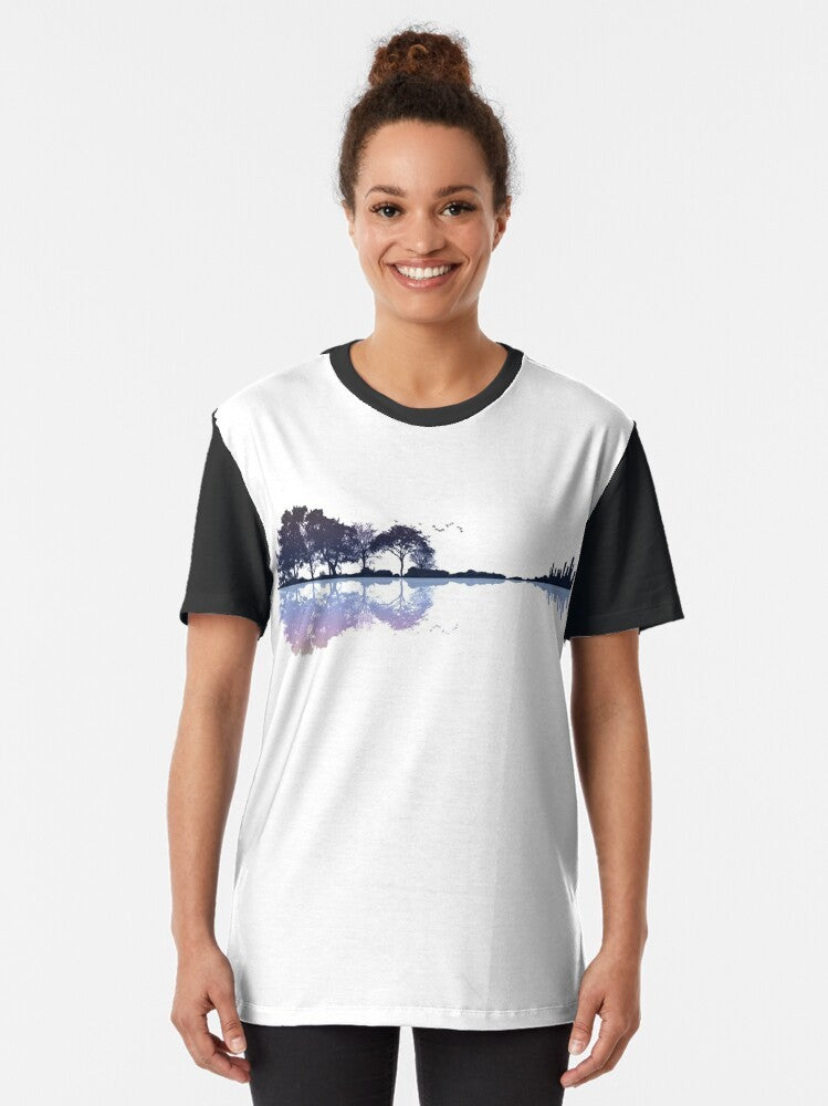 A nature-inspired graphic t-shirt featuring a guitar silhouette against a landscape with trees, water, and a cityscape. - Women