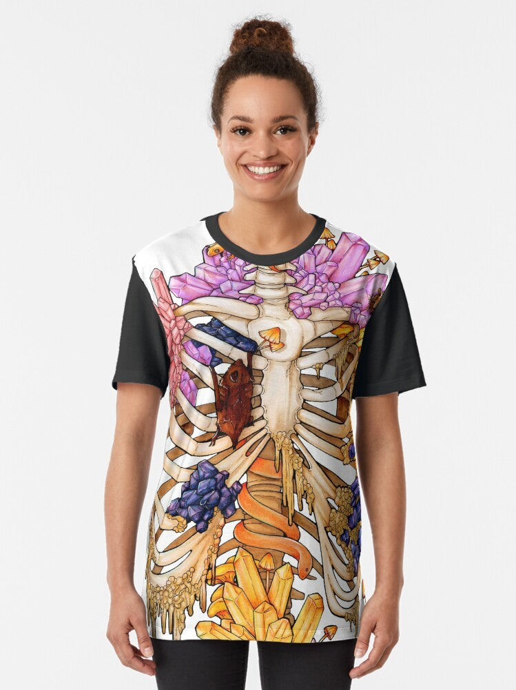 A graphic t-shirt featuring a hidden gem skeleton design in pink and purple colors, surrounded by elements like bats, snakes, mushrooms, and rocks. - Women